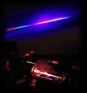 Licht mit hohem UV-Anteil: Die Strahlung, die aus der photonischen Kristallfaser austritt, fächern die Erlanger Forscher mit einem Prisma auf ähnliche Weise auf, wie Wassertropfen Sonnenlicht zu einem Regenbogen brechen. Dabei erhalten sie ein Spektrum (oberer Teil des Bildes), das den sehr breiten ultravioletten Anteil der Strahlung zeigt (rechter Teil des Spektrums). Die normalerweise unsichtbare UV-Strahlung wird durch die Photolumineszenz, die durch das energiereiche UV-Licht induziert wird, sichtbar. An den sichtbaren Bereich (linker Teil des Spektrums) schließt sich der unsichtbare, sehr große infrarote Abschnitt des Spektrums an. (Bild: Xin Jiang)