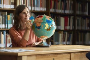 Eine Person sitz vor mehreren Bücherregalen an einem Holztisch und betrachtet einen Globus