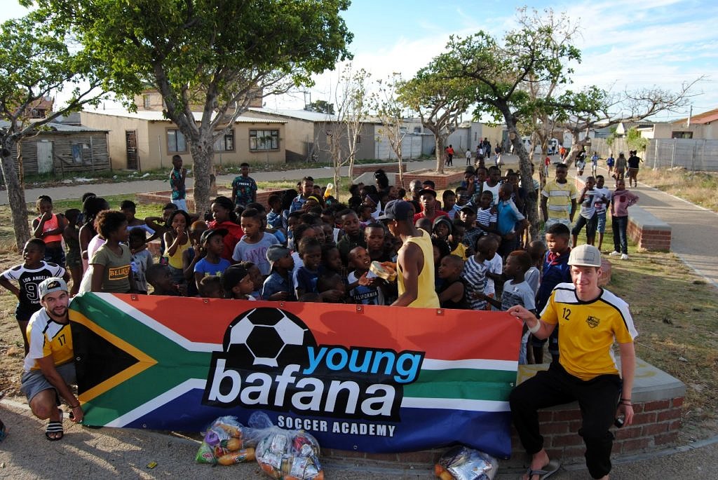 Ehrenamtliche Helfer sind mit den Kindern auf einem Platz versammelt und halten ein Roll-up mit der Aufschrift "Young Bafana Soccer Academy".