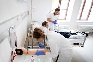 Eine Studentin nimmt an einem künstlichen Arm Blut ab; im Hintergrund behandelt ein Student den Simulationspatienten.