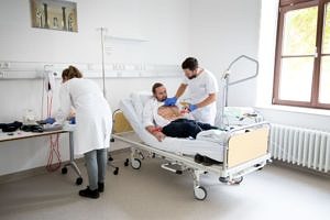 Student behandelt Simulationspatient, von hinten sieht man eine Studentin, die an einem künstlichen Arm das Blutabnehmen übt.