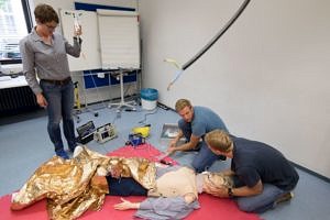 Zwei Studierende knien am Boden und versorgen eine Simulationspuppe. Eine Frau hält den Infusionsbehälter.