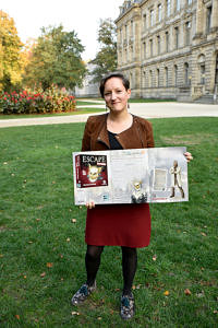FAU-Mitarbeiterin hält im Schlossgarten eine Broschüre zum neuesten Spiel in der Hand.
