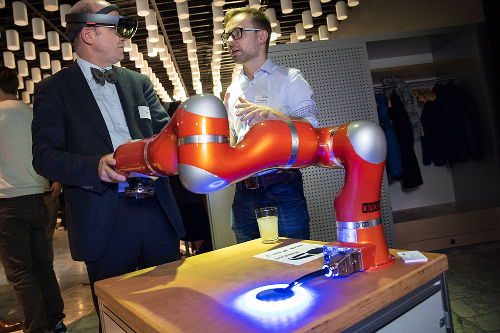 Im Vordergrund ist ein orange-farbener Roboterarm. Im Hintergrund unterhalten sich zwei Männer über dieses Modell. Der eine trägt eine Datenbrille, mit dessen Hilfe Menschen und Roboter miteinander kommunizieren können.