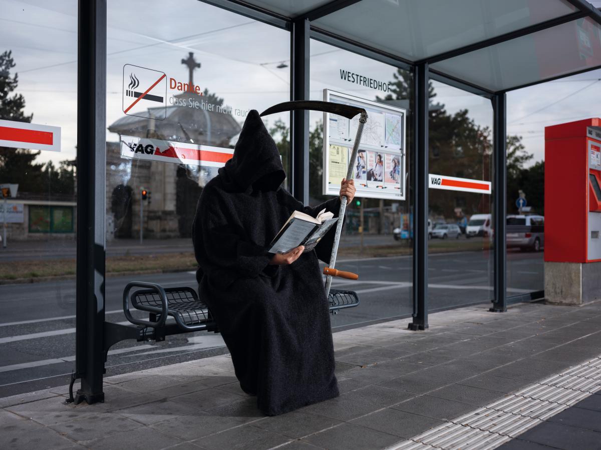 Gevatter Tod liest ein Buch an der Bushaltestelle