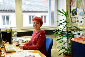 Portrait der Referentin: Sie sitzt an ihrem Arbeitsplatz und lacht in die Kamera. Besonders auffällig sind ihre knall-roten kurzen Haare.
