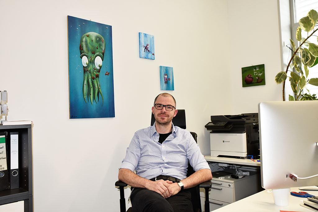 Portraitbild mit Dr. Alexander Scherer an seinem Arbeitsplatz. Im Hintergrund hängen Bilder von Oktopussen.