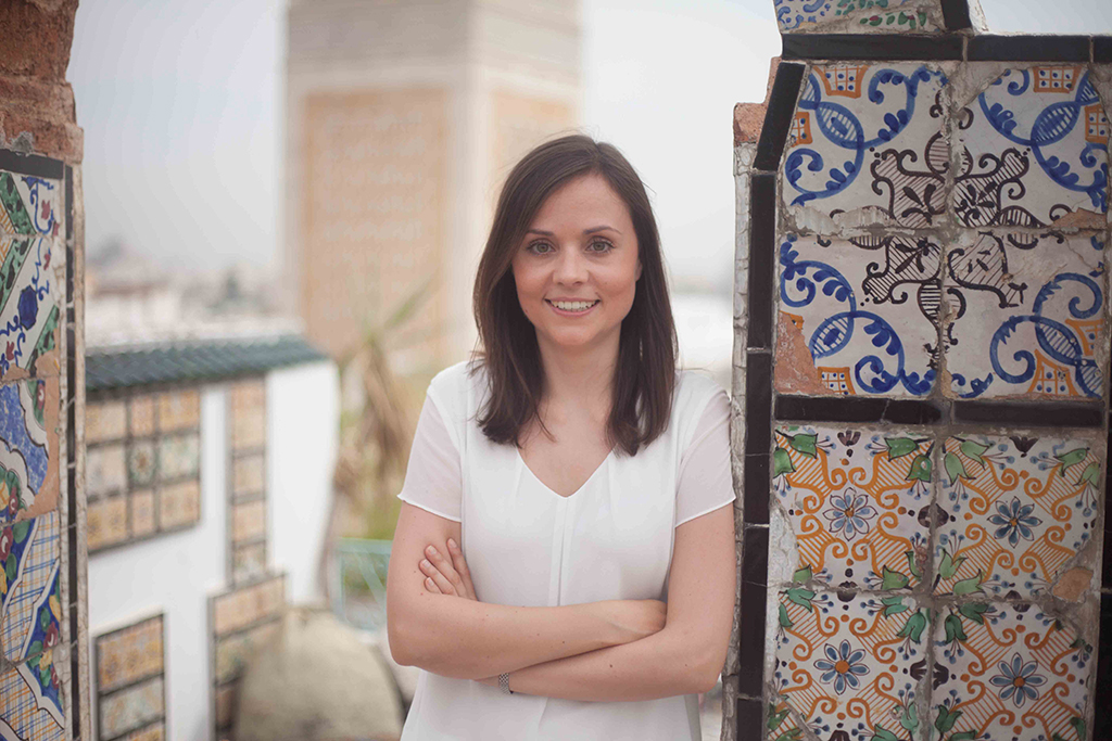 Portraitbild von Johanna Scholz: Sie ist an eine Wand mit bunten Verzierungen gelehnt. Im Hintergrund sieht man noch israelische Gebäude.