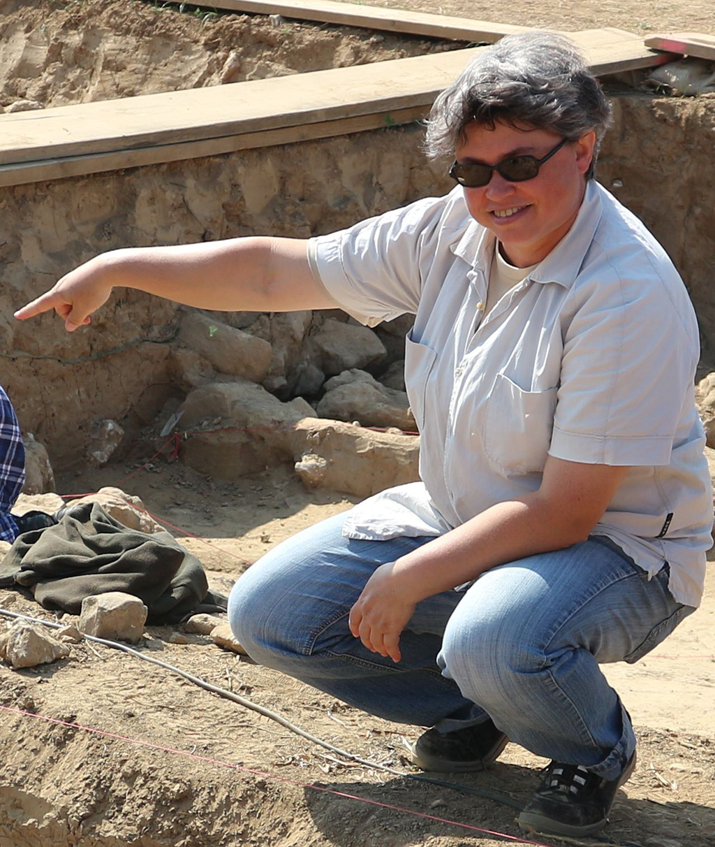 Prof. Mischka an einem Ausgrabungsort: Sie sitzt in der Hocke und zeigt auf etwas. Im Hintergrund ist eine Art Sandgrube zu sehen.