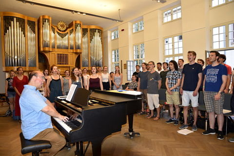 Foto von Chorprobe: Chorleiter ist sitzend am Klavier, Studierende stehen alle. Im Hintergrund eine große Orgel.