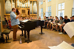 Bild einer Chorprobe: Studierende sitzen und blicken in ihre Liedbücher, während der Chorleiter stehend dirigiert.