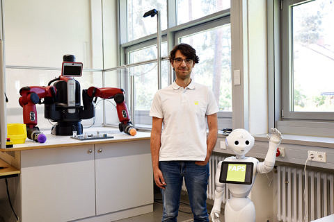Rechts des Mitarbeiters ist ein Roboter mit Gesicht zu sehen, der winkt; links des Mitarbeiters ist ein maschinenartiger Roboter mit zwei Greifarmen zu sehen.