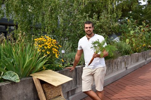 Beschäftigter posiert mit einem Bund Karotten vor einer Grünanlage mit verschiedenen Pflanzen und Blumen.