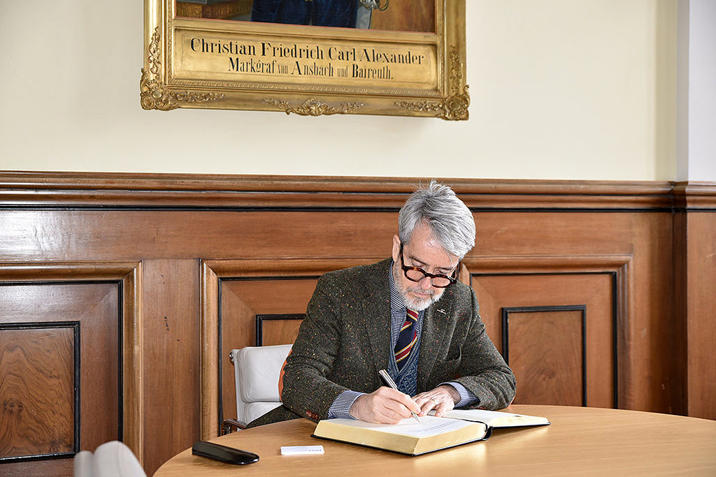Ein Mann mit Brille und grauen Haaren sitzt über einem Buch mit goldenen Seiten und schreibt etwas hinein. Im Hintergrund ist noch ein Teil eines Gemäldes zu sehen.