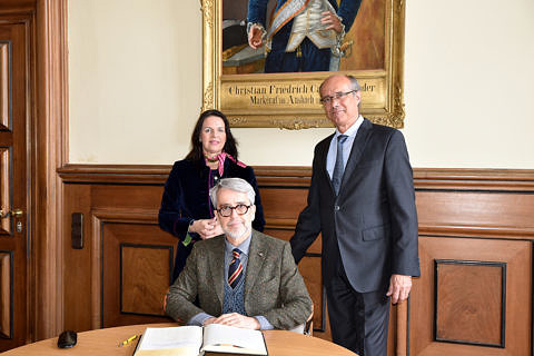 Gruppenbild mit einer Frau (links) und zwei Männern (rechts und in der Mitte sitzend). Vor dem sitzenden Mann liegt ein Buch aufgeschlagen. Im Hintergrund ist ein Teil eines Gemäldes zu sehen.