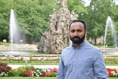 Inder mit Bart und hellblauem Hemd vor einem Brunnen, aus dem Wasser kommt.