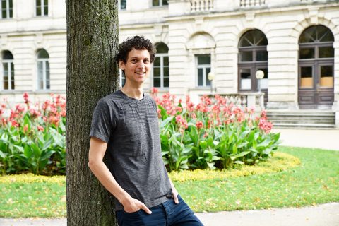 Ein Mann mit grauem Shirt und Jeans steht locker an einem Baum gelehnt und blickt in die Kamera. im Hintergrund ist ein Blumenbeet aus roten Tulpen sowie ein altes Gebäude zu sehen.