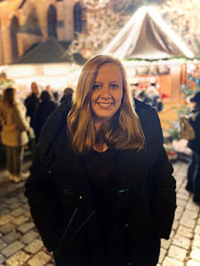 Eine Frau mit langen blonden Haaren steht in schwarzem Mantel auf einem Weihnachtsmarkt und lächelt in die Kamera.