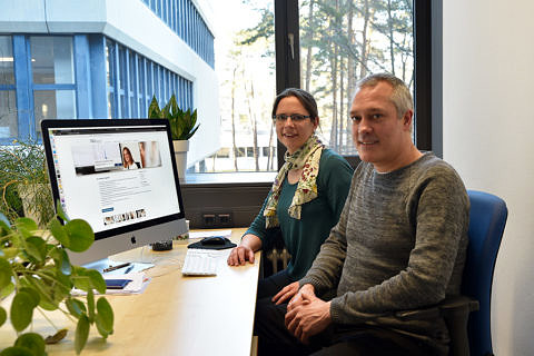 Eine Frau (links) und ein Mann sitzen nebeneinander und Blicken in die Kamera. Im Hintergrund ist der Computerbildschirm mit geöffnetem Browser zu sehen.