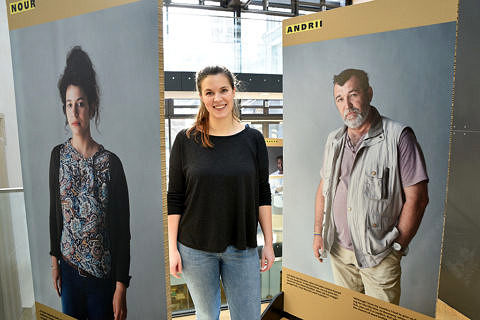 Eine junge Frau mit Jeans, schwarzem Shirt und Pferdeschwanz steht zwischen zwei großen Pappaufstellern, auf denen jeweil ein großes Fotoprorträt von einer Frau (links) und von einem Mann (rechts) abgedruckt sind.