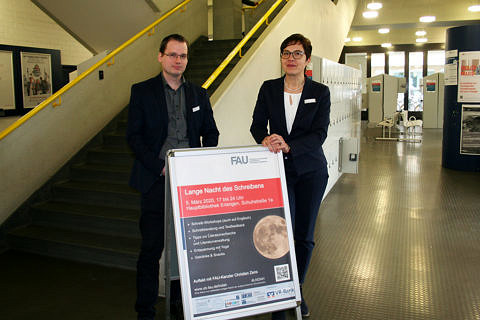 Ein Mann und eine Frau stehen hinter einem Plakataufsteller und blicken in die Kamera.