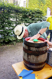 Ein älterer Mann mit Sonnenhut zapft sich Bier aus einem Bierfass.