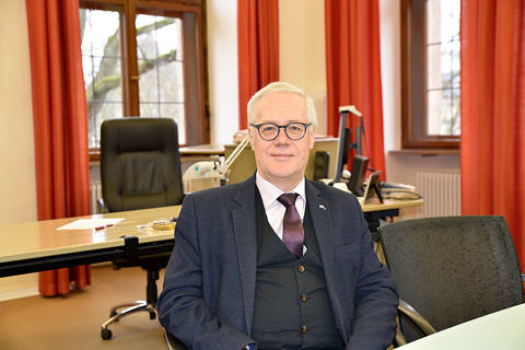 Älterer Herr in Anzug und Krawatte sitzt in einem Büro. Im Hintergrund sind ein Arbeitsschreibtisch sowie rote Vorhänge zu sehen.