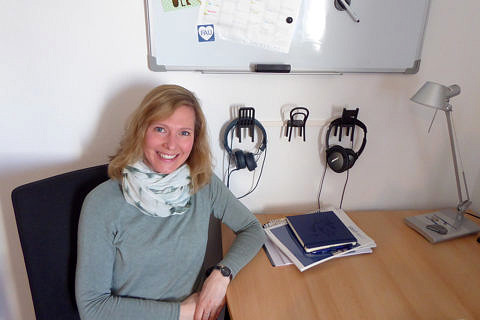 Eine Frau mit blonderen Haaren sitzt an einem Schreibtisch und lächelt in die Kamera. Im Hintergrund hängen Kopfhörer sowie eine Tafel an der Wand.