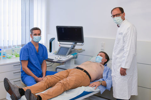 Patient liegt auf einem Bett, zwei Ärzte stehen und sitzen neben ihm.