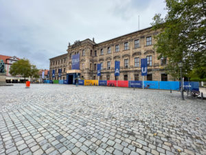 Schlossplatz zeigt Schloss mit Flaggen und Bauzaunbespannung mit dem neuen FAU-Logo