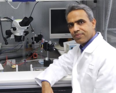 Dr. Bishnoi sitz in einem weißen Kittel vor einem Mikroskop und blickt in die Kamera.