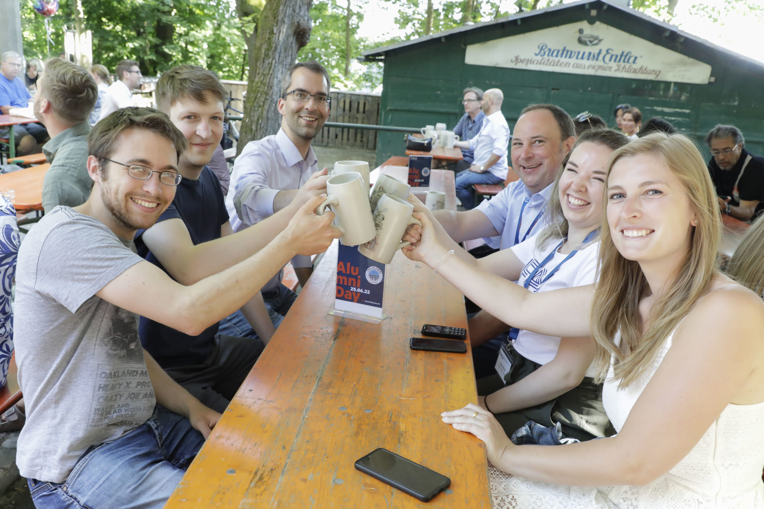 A group of people sitting on benches in the Entlas Biergaten, clinking tankards and looking at the camera