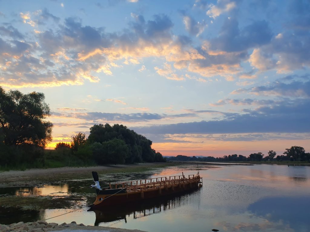 An einem Strand liegt das Römerboot. Im Hintergrund geht die Sonne unter, es ist leicht bewölkt.