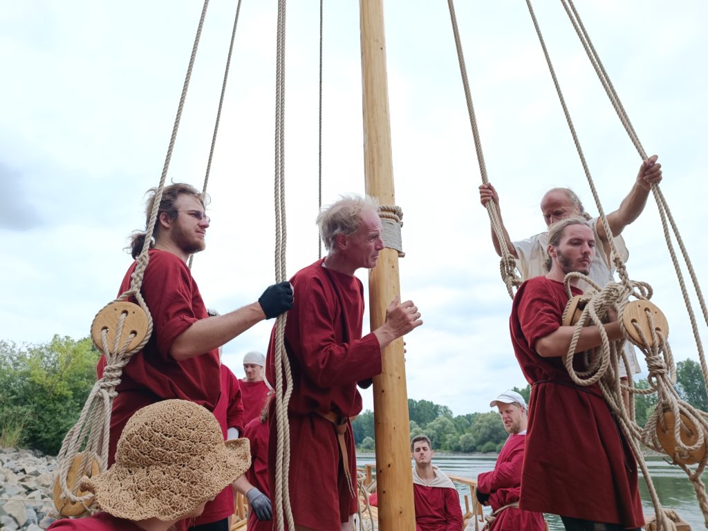 Mehrere Männer in roten und weißen Gewändern stehen auf einem Römerboot.