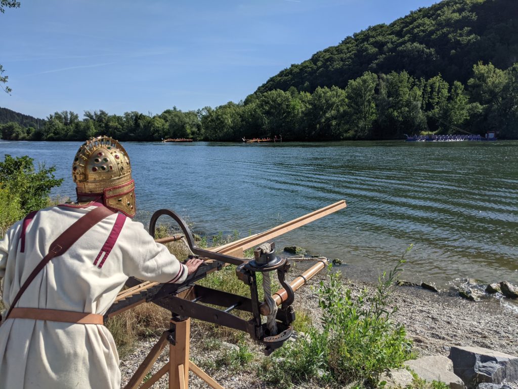 An einem Geschütz steht ein Mann in römischer Soldatenuniform und blickt auf den Fluss hinauf.