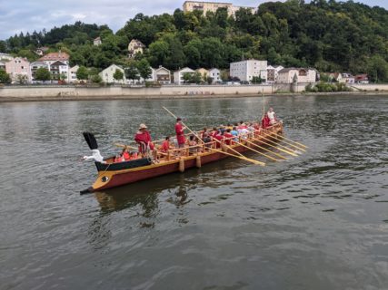 Ein Römerboot schwimmt auf der Donau. Im Boot sitzen Touristen und Crewmitglieder in roten Klamotten.