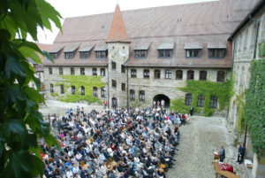 In einem Innenhof sitzen viele Leute auf Stühlen. Vorne am Rednerpult steht ein Mann an einem Mikrofon, daneben ist ein Tisch aufgebaut.