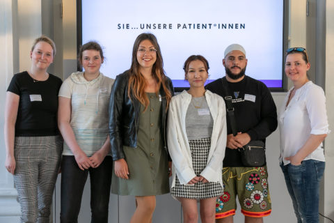 Ein Gruppenbild von sechs Personen: fünf Frauen und ein Mann stehen nebeneinander und lächeln in die Kamera. Hinter Ihnen steht auf einer Leinwand: "Sie- unsere Patient*innen".