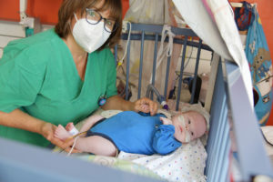 Eine Pflegerin in grünen Klamotten steht am Krankenbett eines Babys und schaut in die Kamera. DEr Junge wird beatmet und umklammert den Daumen der Pflegerin.
