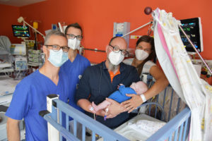 Zwei Ärzte in blauen Arztklamotten stehen neben den Eltern in einem Krankenzimmer. Der Vater hält das kleine Kind, die Mutter steht hinter ihm und streichelt den Rücken des Babys.