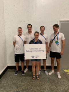 Fünf Personen stehen vor einer weißen Wand und halten ein Schild mit der Schrift "University of Erlangen-Nürnberg".