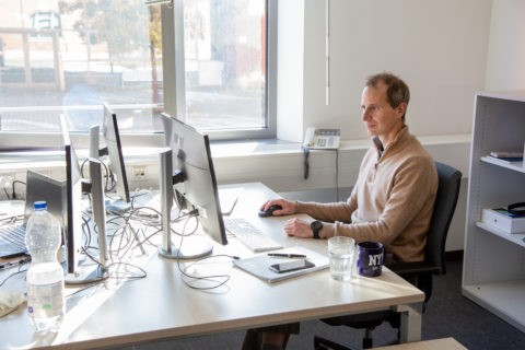 Ein Mann in beigem Pullover sitzt am Schreibtisch vor dem Computer.