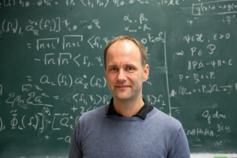 Ein Mann im Pullover steht vor einer Tafel mit mathematischen Gleichungen.