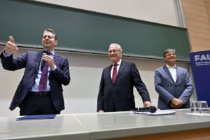 Drei Männer in Anzügen vor einer großen Tafel in einem Hörsaal. Der Mann links hält ein Mikro in der Hand.