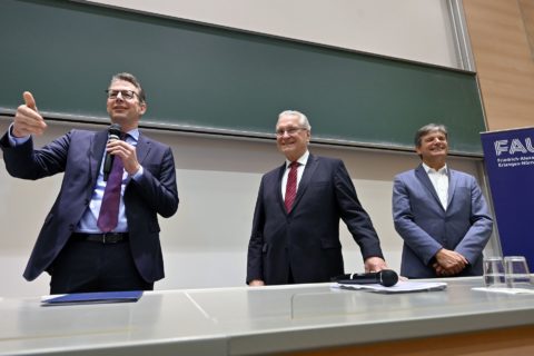 Drei Männer in Anzügen vor einer großen Tafel in einem Hörsaal. Der Mann links hält ein Mikro in der Hand.