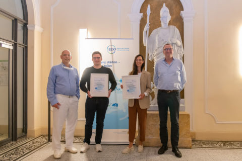 Gruppenbild: In der Mitte stehen eine Frau und ein Mann, beide halten eine Urkunde hoch. Links und rechts von ihnen stehen zwei Männer.