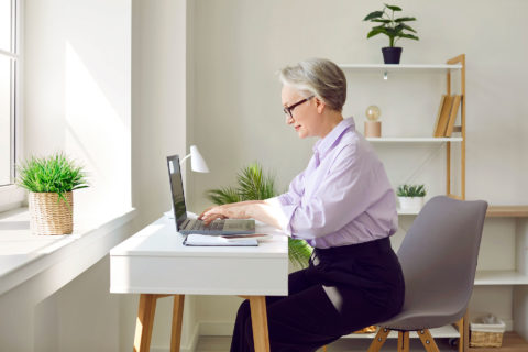 Eine Frau mit grauen Haaren sitzt an einem Schreibtisch am Laptop.