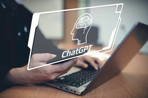 Mensch an Laptop, Ausschnitt auf Hände und Laptop, Darüber liegt Illustration von Kopf mit Gehorn und dem Text ChatGPT