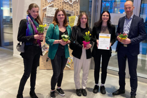 Vier Frauen und ein Mann posieren mit Blumen und einer Urkunde in einer Reihe.