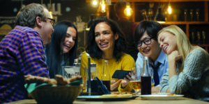 Frau zeigt ihren Freunden interessante Sachen auf ihrem Smartphone, während sie in der Bar gute Zeit haben. Sie lachen, scherzen, trinken in Hipster-Bar.